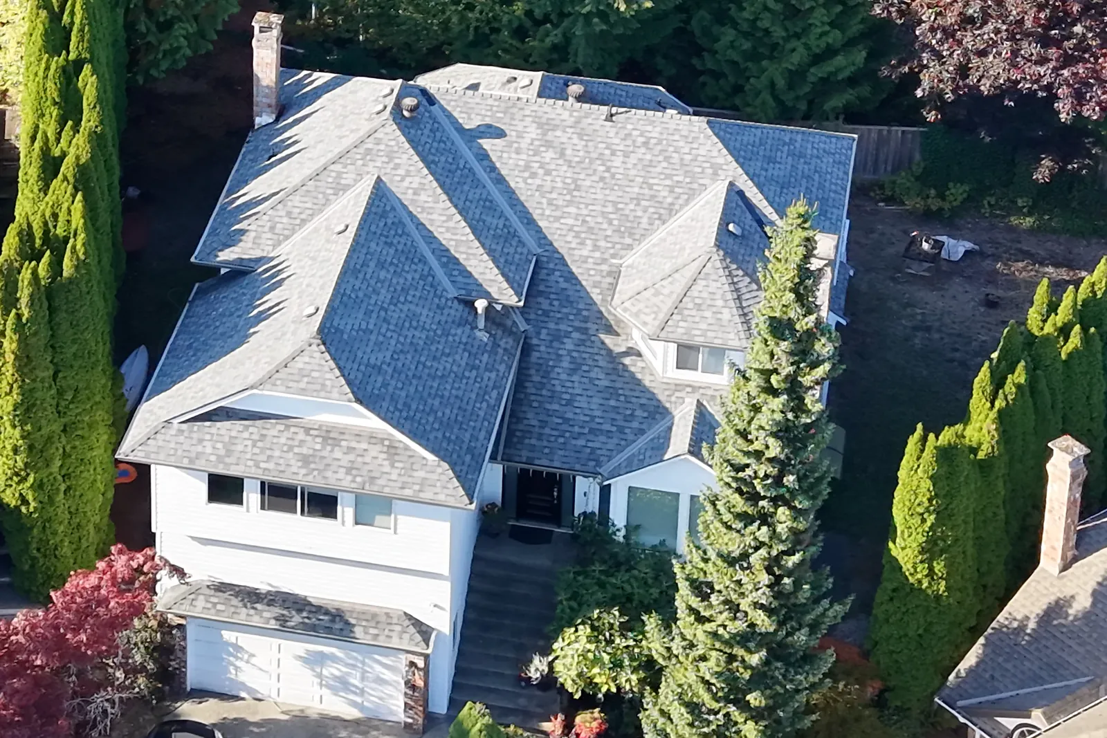 Looking down on a white two-storey house with a grey shingle roof, front steps and garden, surrounded by tall evergreens
