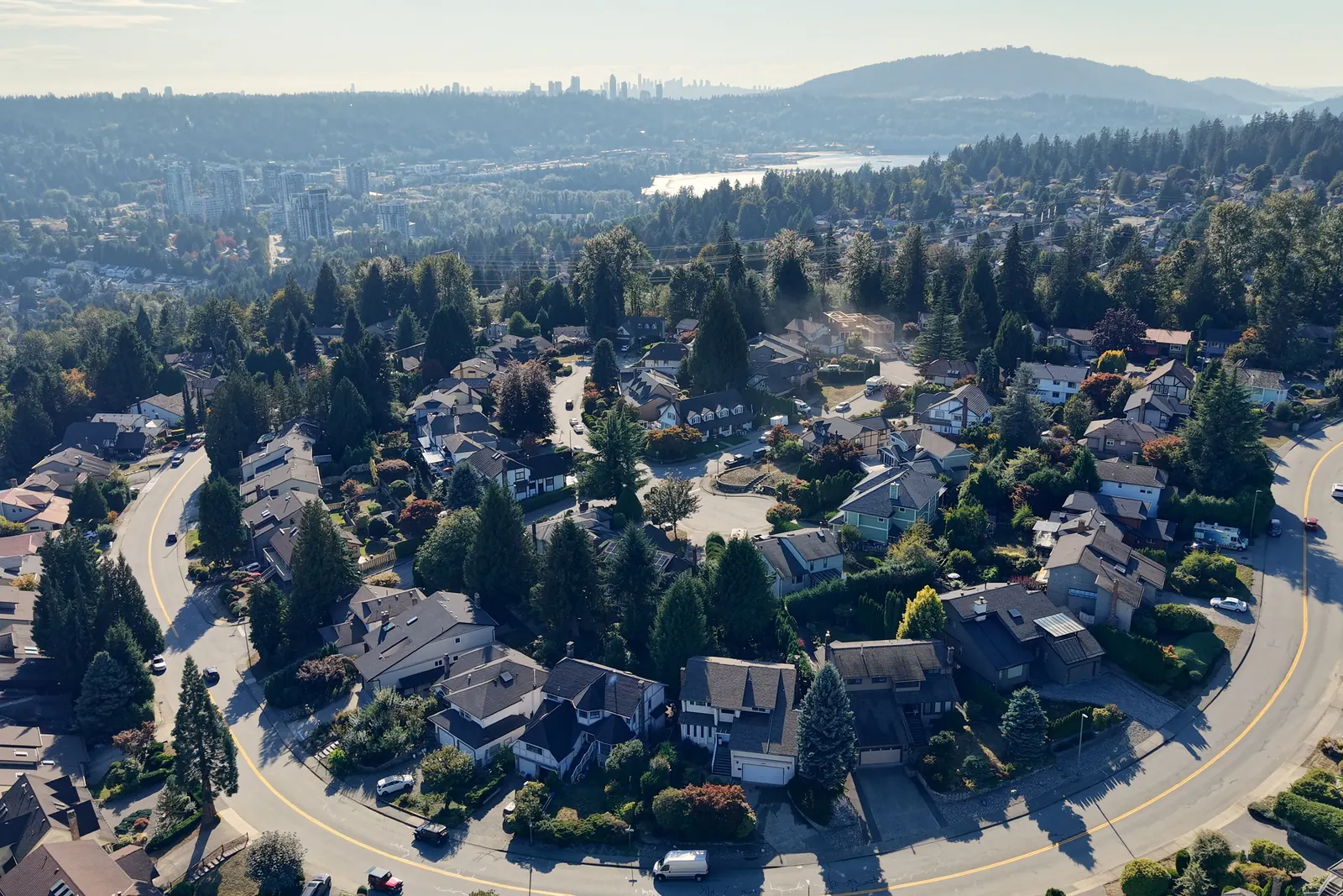 A curving road through a neighbourhood of houses and tall evergreens, with water, hills and distant towers on the horizon